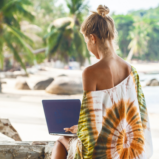 Frau sitzt entspannt am Strand auf einer Decke, arbeitet mit einem Laptop, lächelt und genießt die Sonne. Symbol für Freiheit, Balance und selbstbestimmtes Arbeiten als berufstätige Mutter.