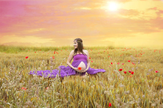 Schwangere Frau sitzt in einer Wiese mit Mohn in einem Lila Kleid