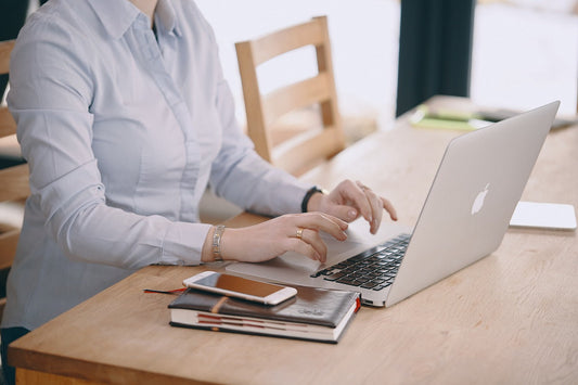 Oberkörper einer Frau ohne sichtbaren Kopf, die an einem Tisch sitzt und mit Laptop, Terminplaner und Smartphone arbeitet – Symbol für Online-Business als Mutter.